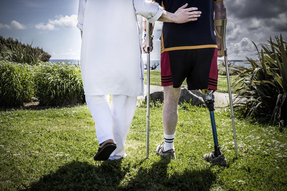 A person in white walks with a man using crutches and a prosthetic leg on a grassy path. The sky is partly cloudy, with a calm ocean in the background. Compassionate tone.