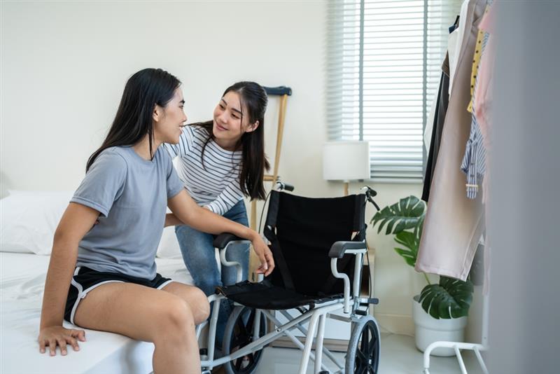 Two women share a warm moment in a bedroom. One is seated on a bed, and the other leans in beside a wheelchair, conveying care and companionship.
