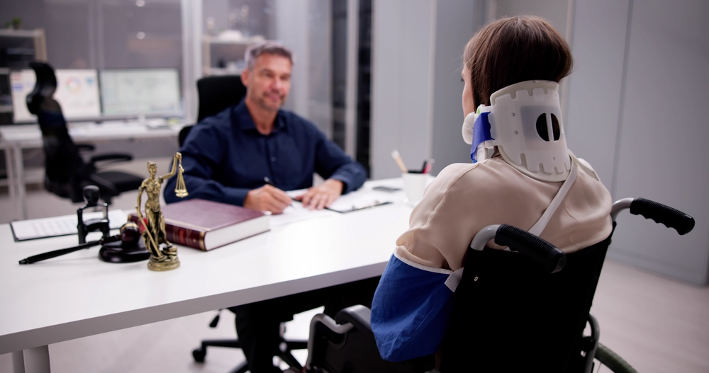 A person in a wheelchair with a neck brace and arm cast consults with a man at a desk. A scale of justice and law book suggest legal advice.