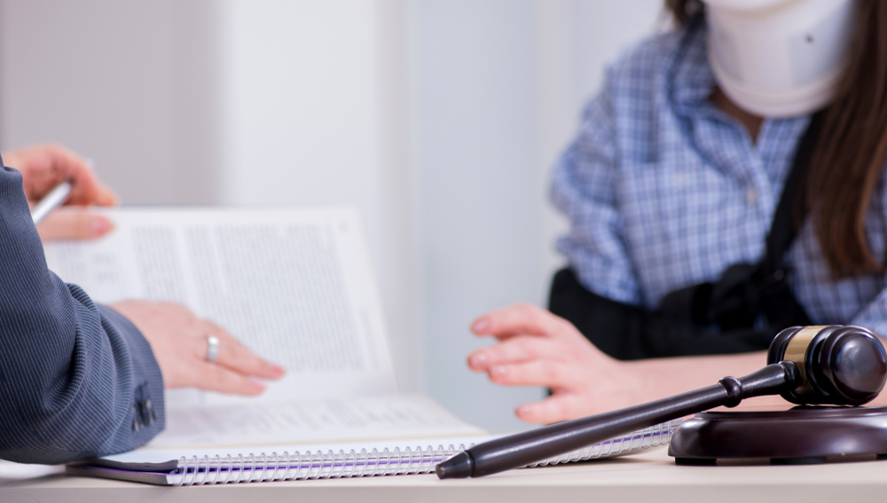 A person in a neck brace and arm sling consults a person holding a document. A gavel rests on a table, suggesting a legal or medical consultation.