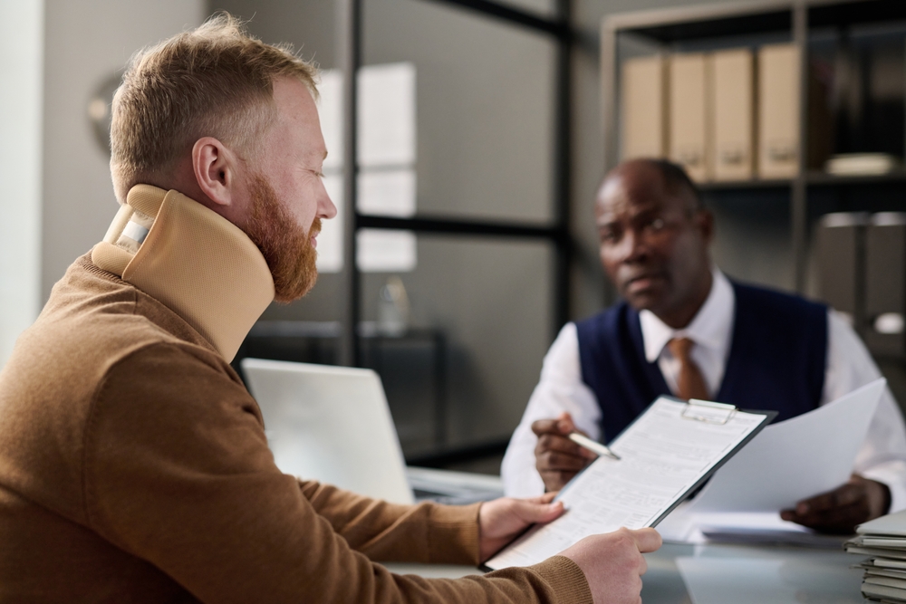A man with a neck brace consults with a professional holding a clipboard. They are in an office setting, discussing documents with focused expressions.