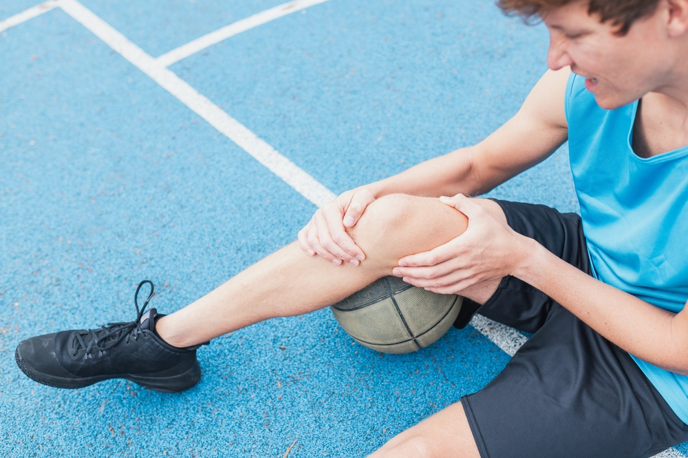 A young person in a blue jersey sits on a blue sports court, holding their knee in pain with a basketball underneath. The scene conveys discomfort and injury.