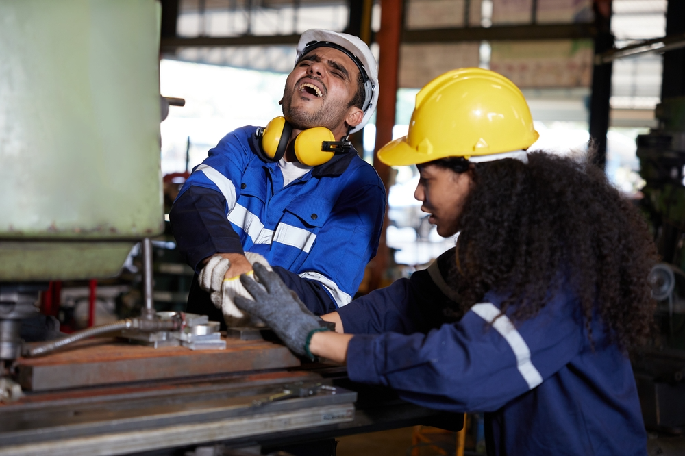 A factory worker in a blue uniform holds his injured hand in pain, while a female colleague in a yellow helmet assists him, conveying urgency.