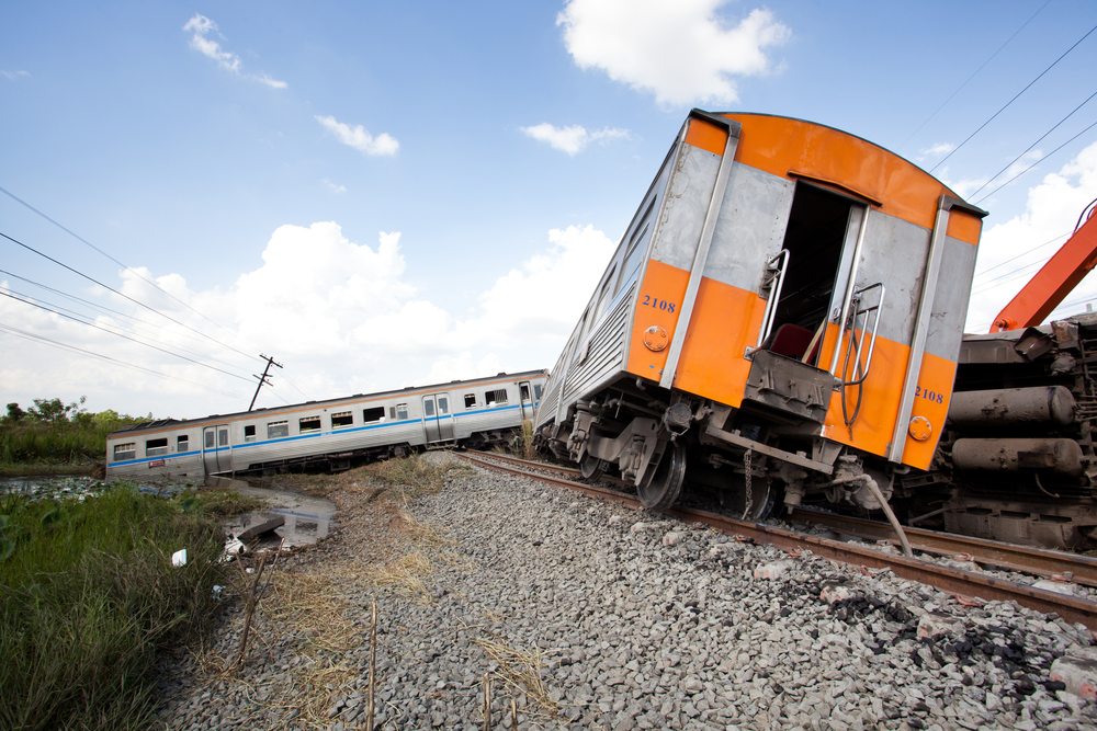 Train derailed on rocky tracks under a clear blue sky. Two carriages tilted, with one partially off the rails. Grassy area and power lines nearby.