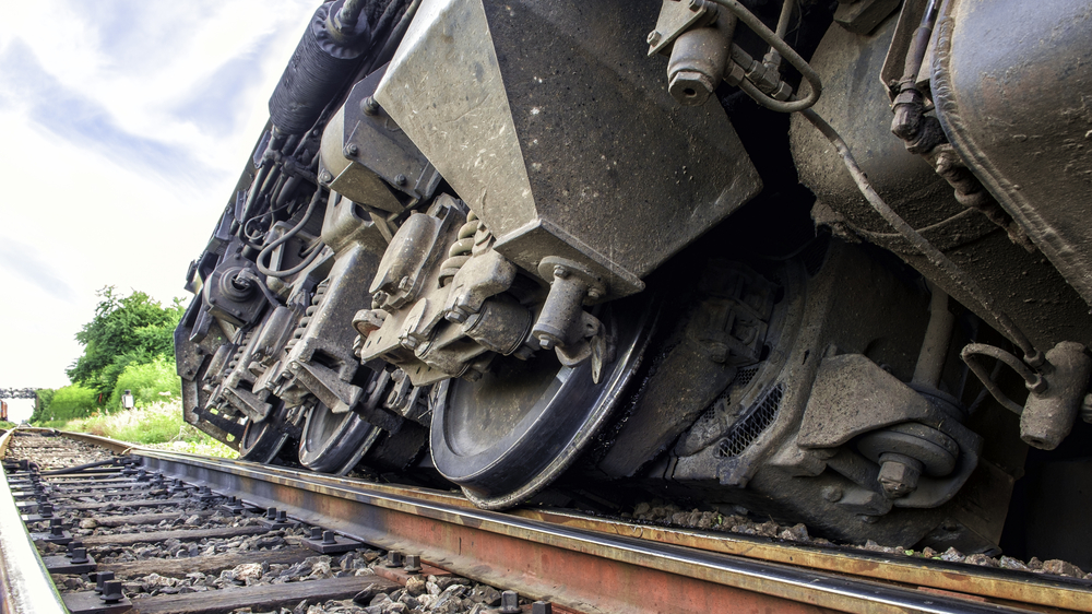 Close-up of a derailed train on its side, showing large metal wheels and mechanisms. The tracks are visible, surrounded by greenery and a cloudy sky.