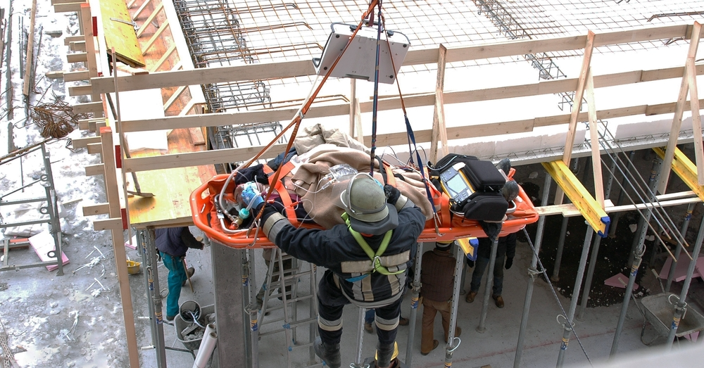 A construction worker in a helmet is rescued using a stretcher and crane on a snowy construction site. Emergency equipment is visible, conveying urgency.