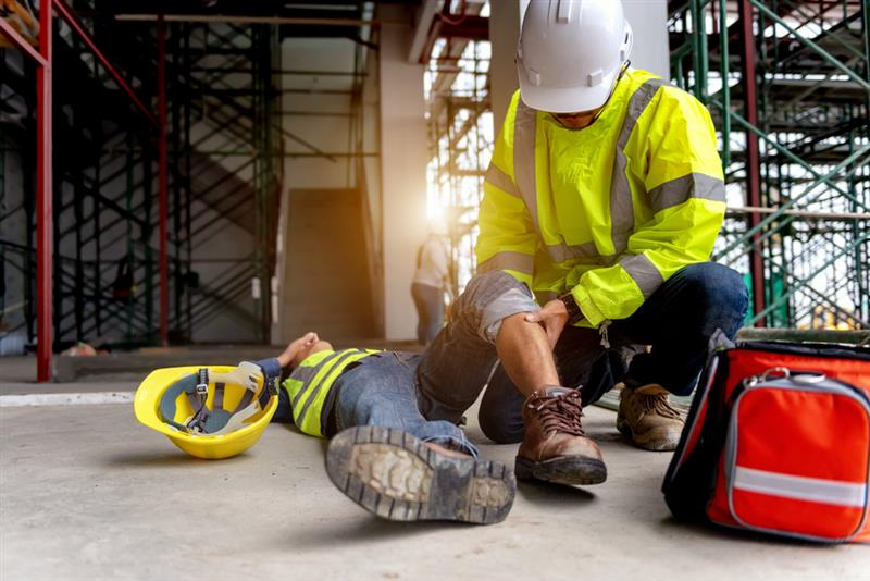 A construction worker in a reflective jacket aids a fallen colleague inside a site. A hard hat lies on the ground beside them, creating a tense atmosphere.