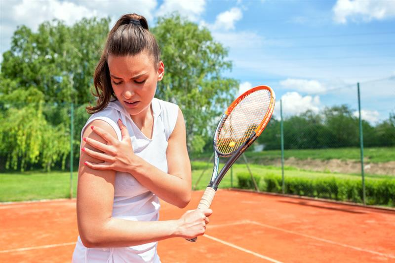 A female tennis player in white holds her shoulder in pain on a clay court. The sky is clear with a few clouds, and lush greenery surrounds the court.