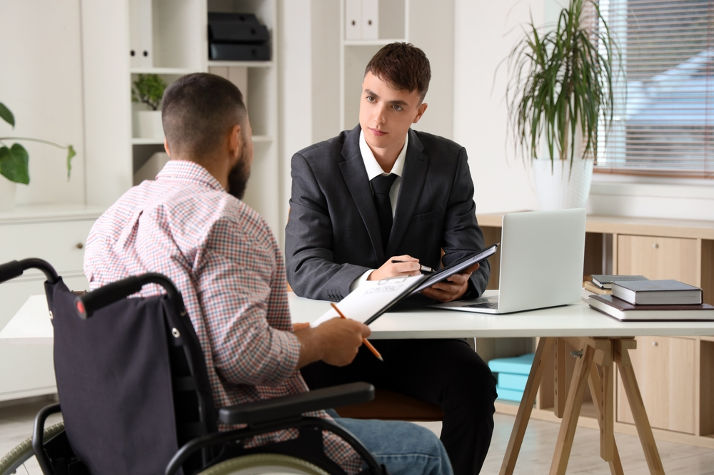 A man in a wheelchair discusses paperwork with a man in a suit. They are in an office setting, fostering a professional and focused atmosphere.