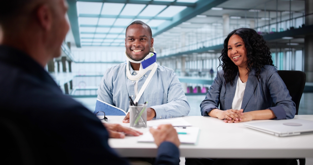 A man with a neck brace and arm sling sits smiling across a table from another person, with a woman beside him. The setting appears to be a modern office.