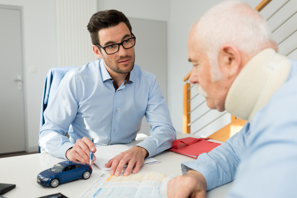 A man in glasses sits at a desk discussing paperwork with an older man wearing a neck brace. A model car is on the table, suggesting a car accident claim.