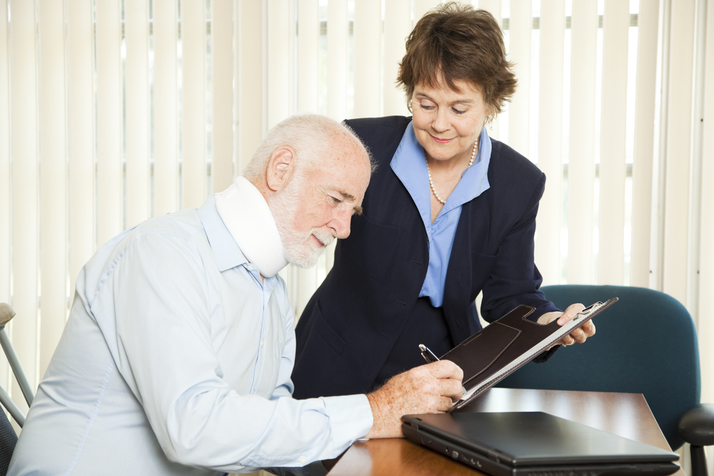 An elderly man with a neck brace signs a document on a clipboard held by a smiling woman in a suit, sitting at a table with a laptop.
