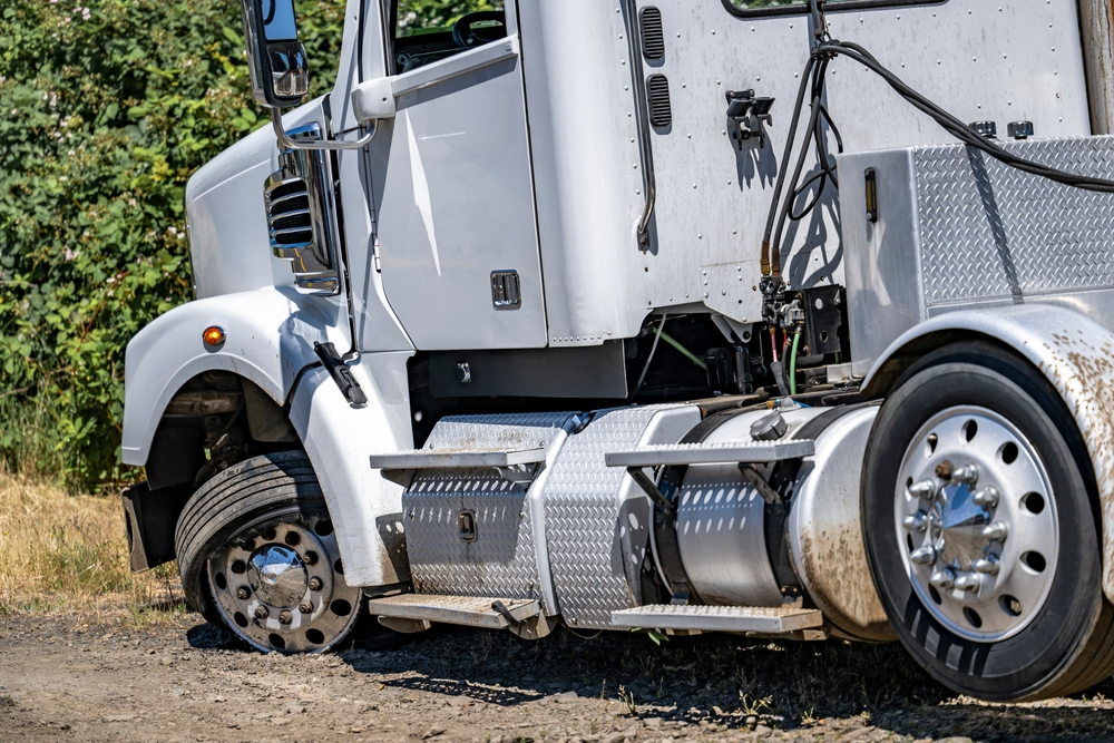 A white semi-truck with a flat rear tire is parked on a rural dirt path. Surrounding bushes and undergrowth suggest a countryside setting, evoking strain and challenge.