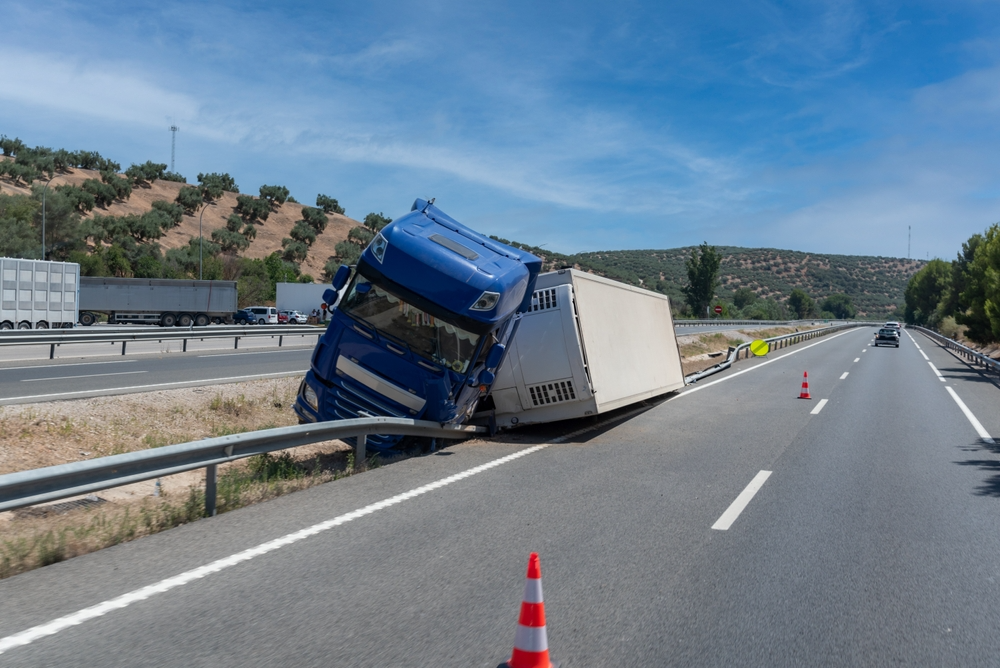 A blue semi-truck is tipped over on the highway, leaning against a barrier. Traffic cones are placed nearby. The sky is clear, and hills are in the background.