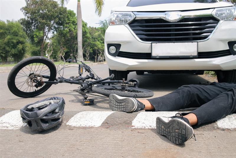 A bicycle and helmet lie on the road next to a person and a white car, suggesting a collision or accident scene.