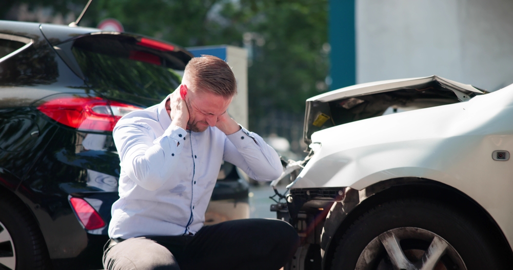 A man in a white shirt crouches near two cars with damaged front ends after a collision, holding his head in distress.