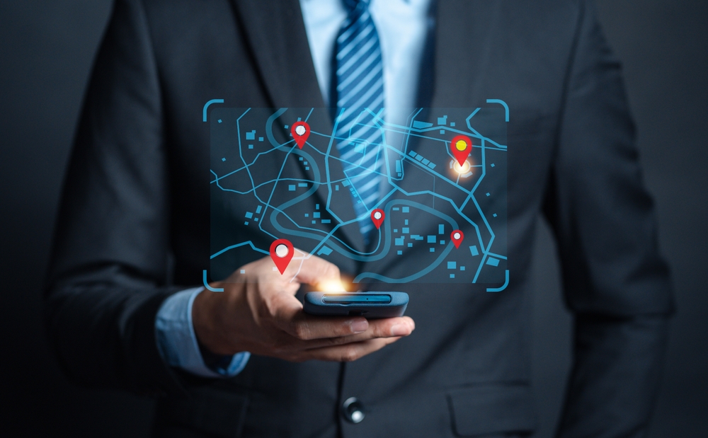 A man in a suit holds a smartphone displaying a digital map with red location pins. The map appears as a glowing hologram, suggesting navigation or GPS use.