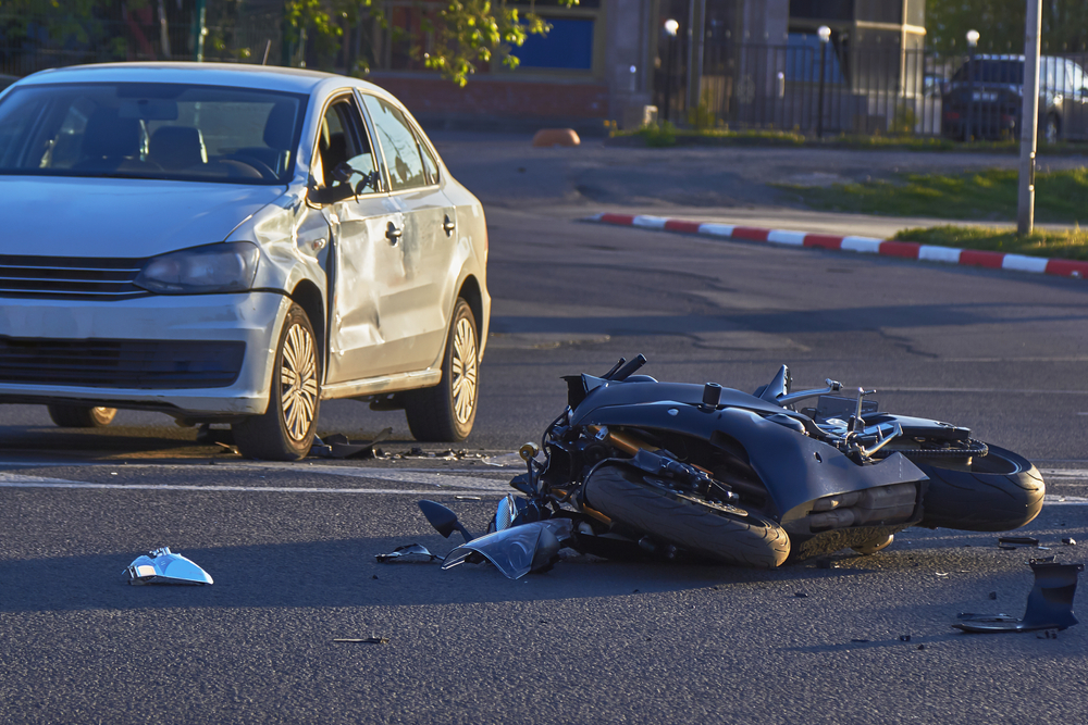A damaged silver car and overturned motorcycle lie on a street after a collision, with debris scattered around them, conveying a sense of urgency and impact.