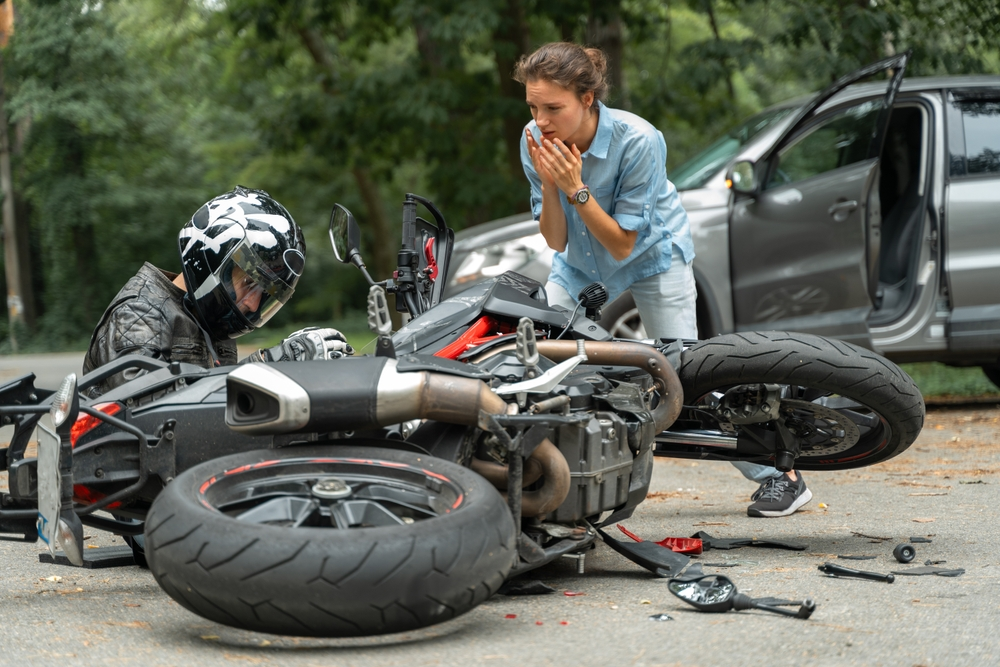 A woman looks worriedly at a motorcyclist sitting near a fallen bike after an accident. A car with an open door is seen in the background, suggesting a collision.