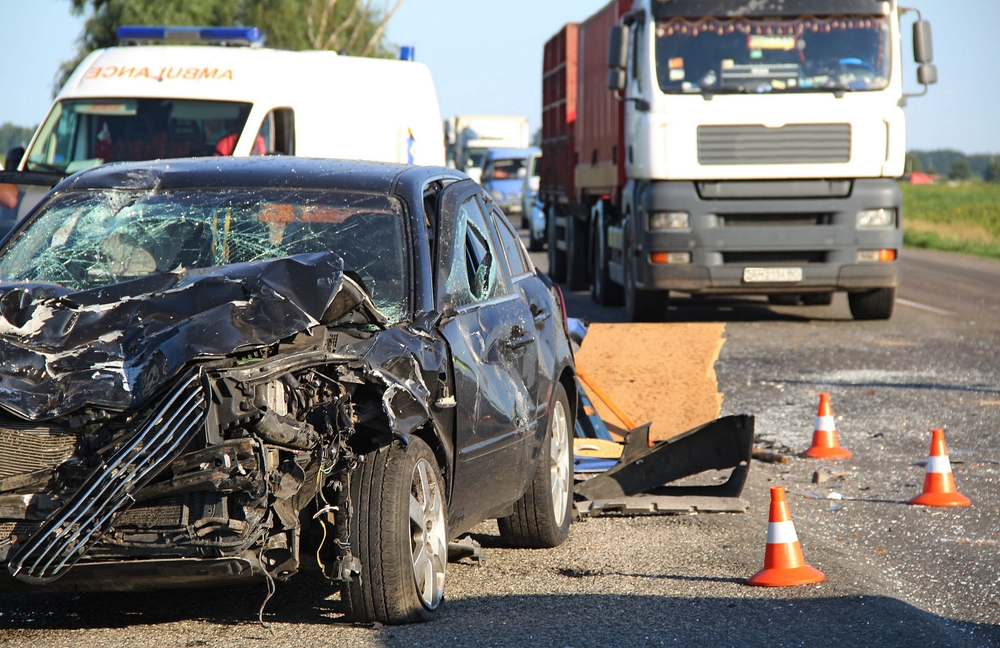 Crashed black car with a severely damaged front on a road. Nearby, an ambulance and a truck are present.
