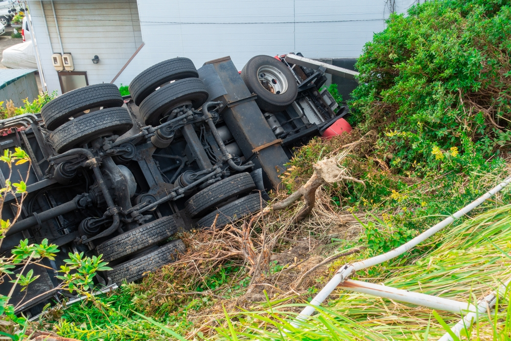 Overturned truck lies on its side amidst bushes and grass by a building, conveying a sense of chaos and disruption in a natural setting.