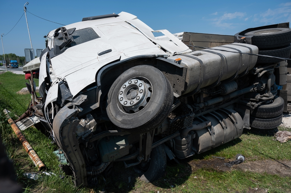 Overturned white truck on grassy roadside, with visible damage and debris. Bright blue sky suggests daytime. Scene implies an accident.