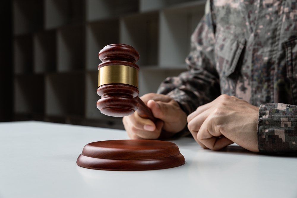A person in military uniform strikes a wooden gavel on a sound block, symbolizing authority and judgement, set against a blurred background.