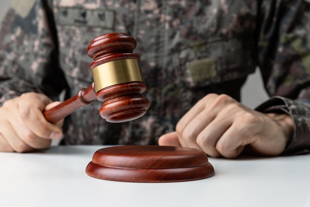 A person in a camouflage uniform holds a wooden gavel over a striking block, conveying authority and decision-making in a military context.