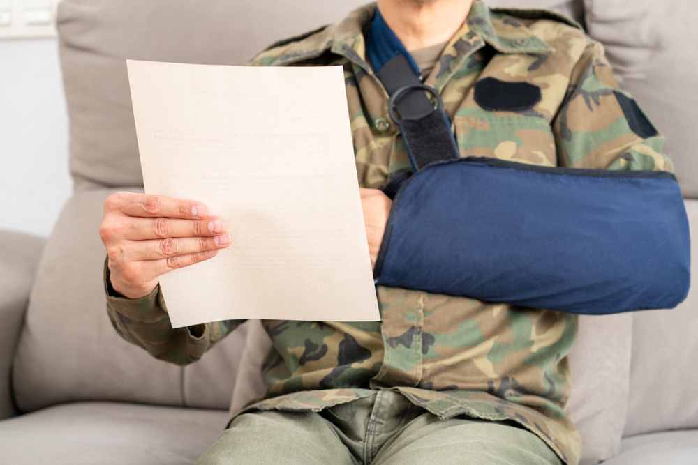 A person in a camouflage uniform with an arm in a navy blue sling sits on a beige couch, holding and reading a paper document, suggesting focus.