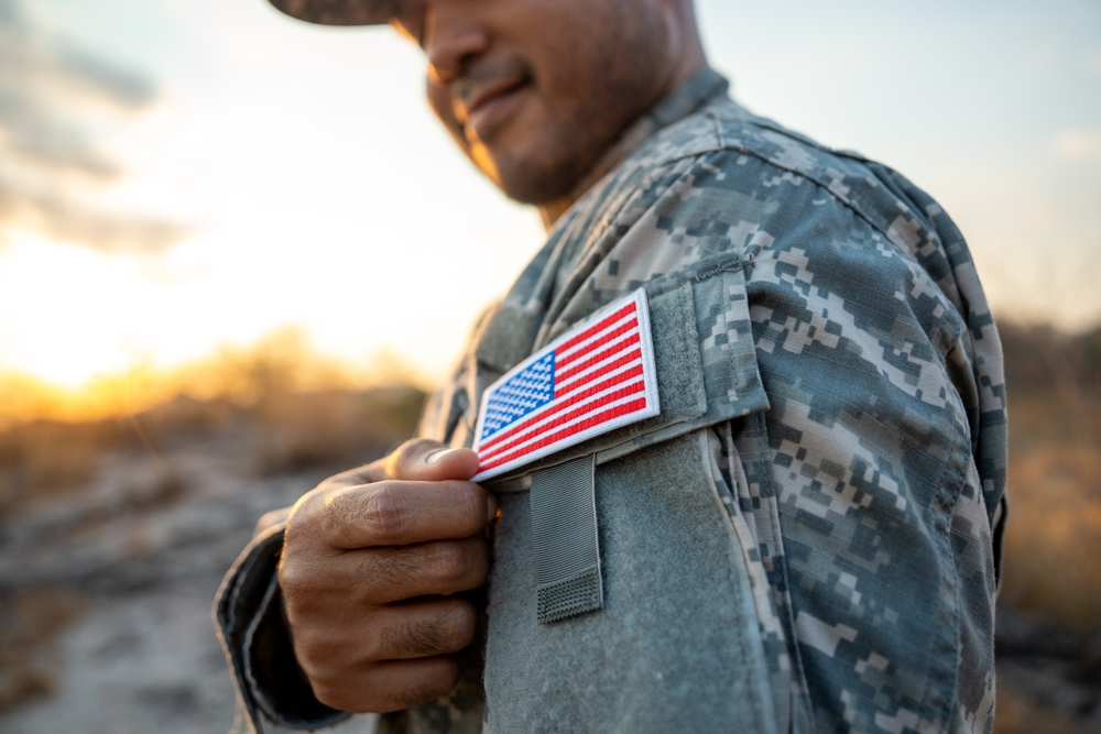 Close-up of a U.S. military service member in camouflage uniform holding an American flag patch on their sleeve during sunset.